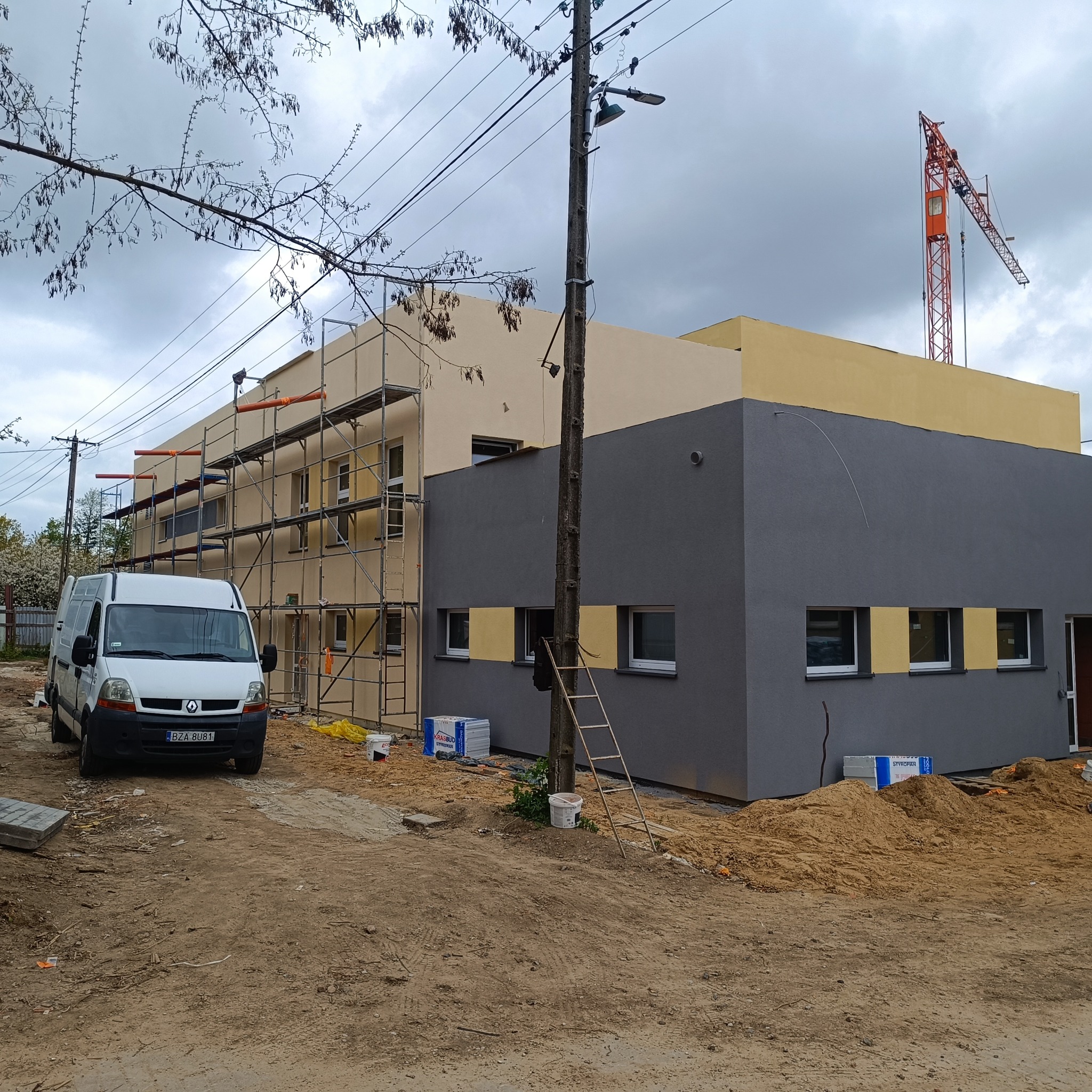 Budynek po renowacji elewacji, z rusztowaniami i zaparkowanym busem. Widoczne świeżo pomalowane fragmenty w odcieniach szarości i beżu. W tle żuraw budowlany.