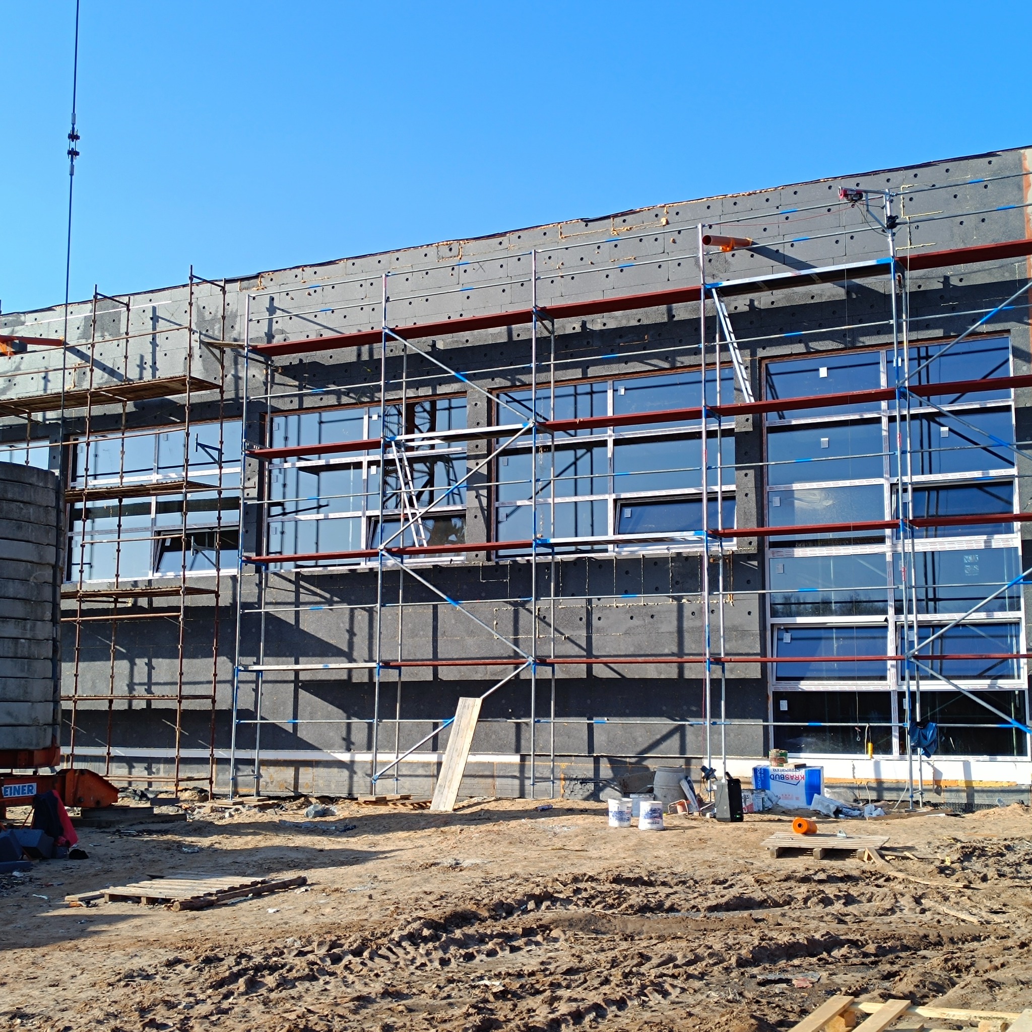 Budynek w trakcie ocieplania, widoczne rusztowania i materiały izolacyjne. Okna zamontowane, teren budowy z ziemią i paletami. Słoneczny dzień.