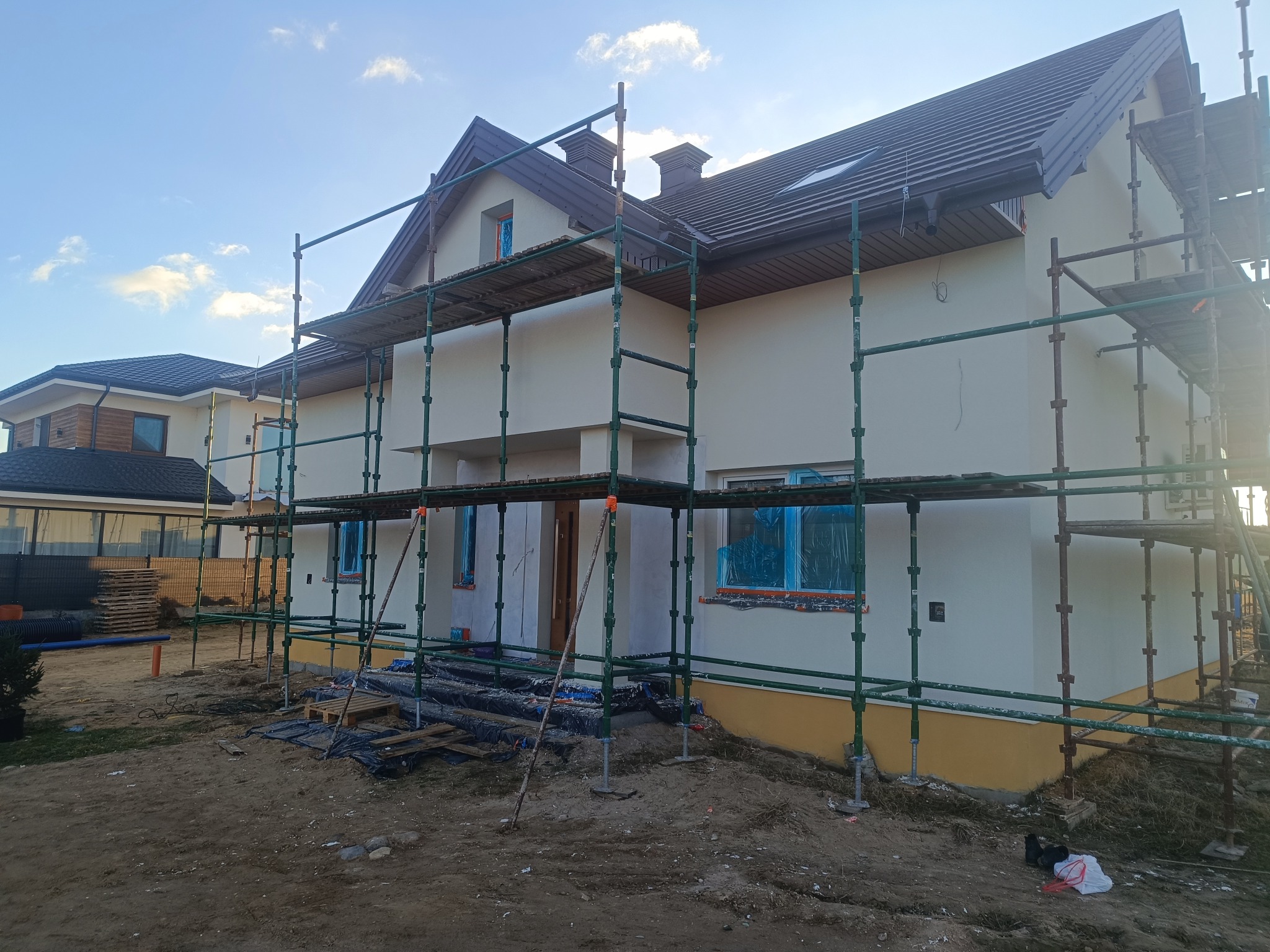 Dom jednorodzinny w trakcie prac elewacyjnych, z widocznymi rusztowaniami i zabezpieczonymi oknami. Świeżo otynkowana fasada w jasnym odcieniu, przygotowana do dalszych prac wykończeniowych.