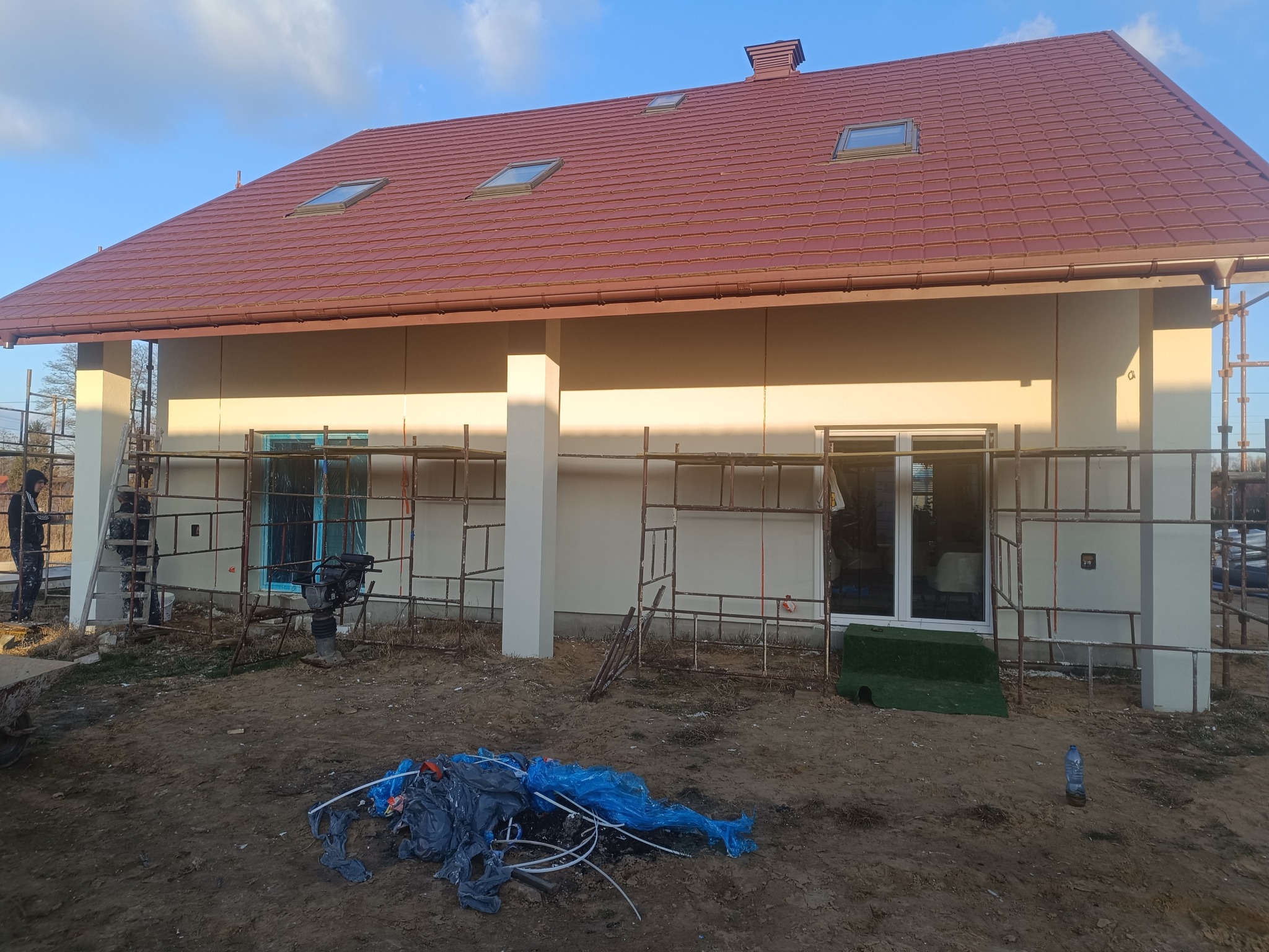 Dom jednorodzinny z beżową elewacją i brązowym dachem z lukarnami, w trakcie prac wykończeniowych. Widoczne rusztowania i materiały budowlane.