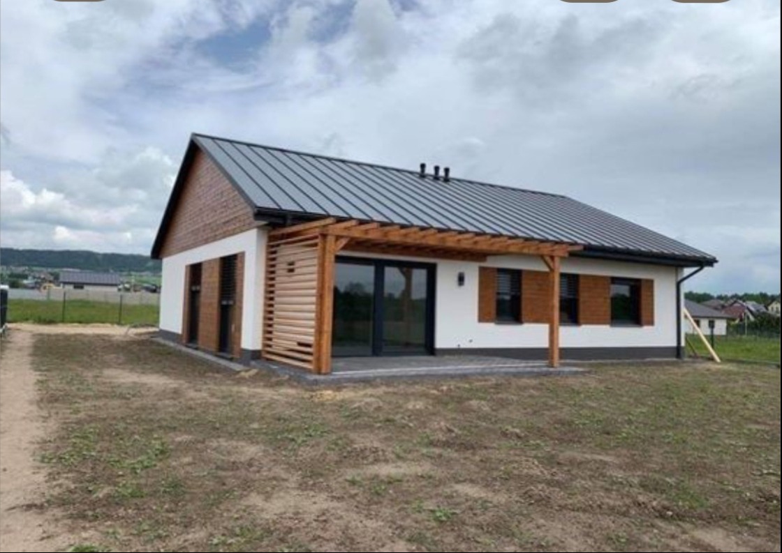 Nowoczesny dom jednorodzinny z białą elewacją, drewnianymi elementami dekoracyjnymi i szarą blachodachówką. Widoczna pergola nad tarasem i starannie wykończone detale.