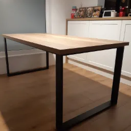 Stół z drewnianym blatem i czarnymi, metalowymi nogami o kwadratowym przekroju, ustawiony na drewnianej podłodze, w tle biała szafka i szara ściana.