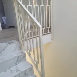 Biała, geometryczna balustrada schodowa z profilami prostokątnymi, zamontowana przy marmurowych schodach w jasnym wnętrzu.