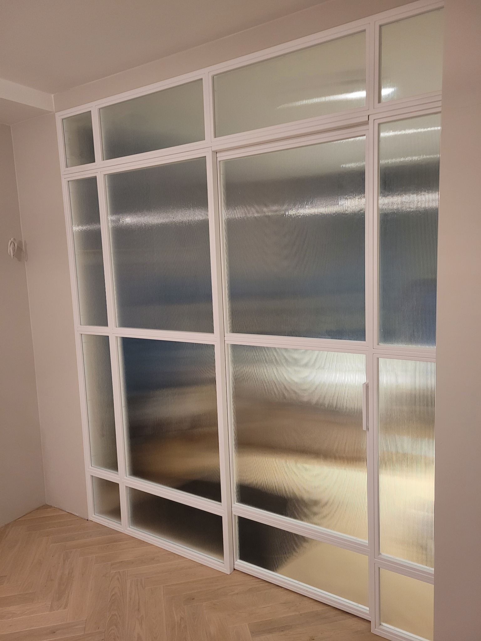 Nowoczesna ścianka działowa z białymi profilami i ryflowanym szkłem, widok na parkiet ułożony w jodełkę.