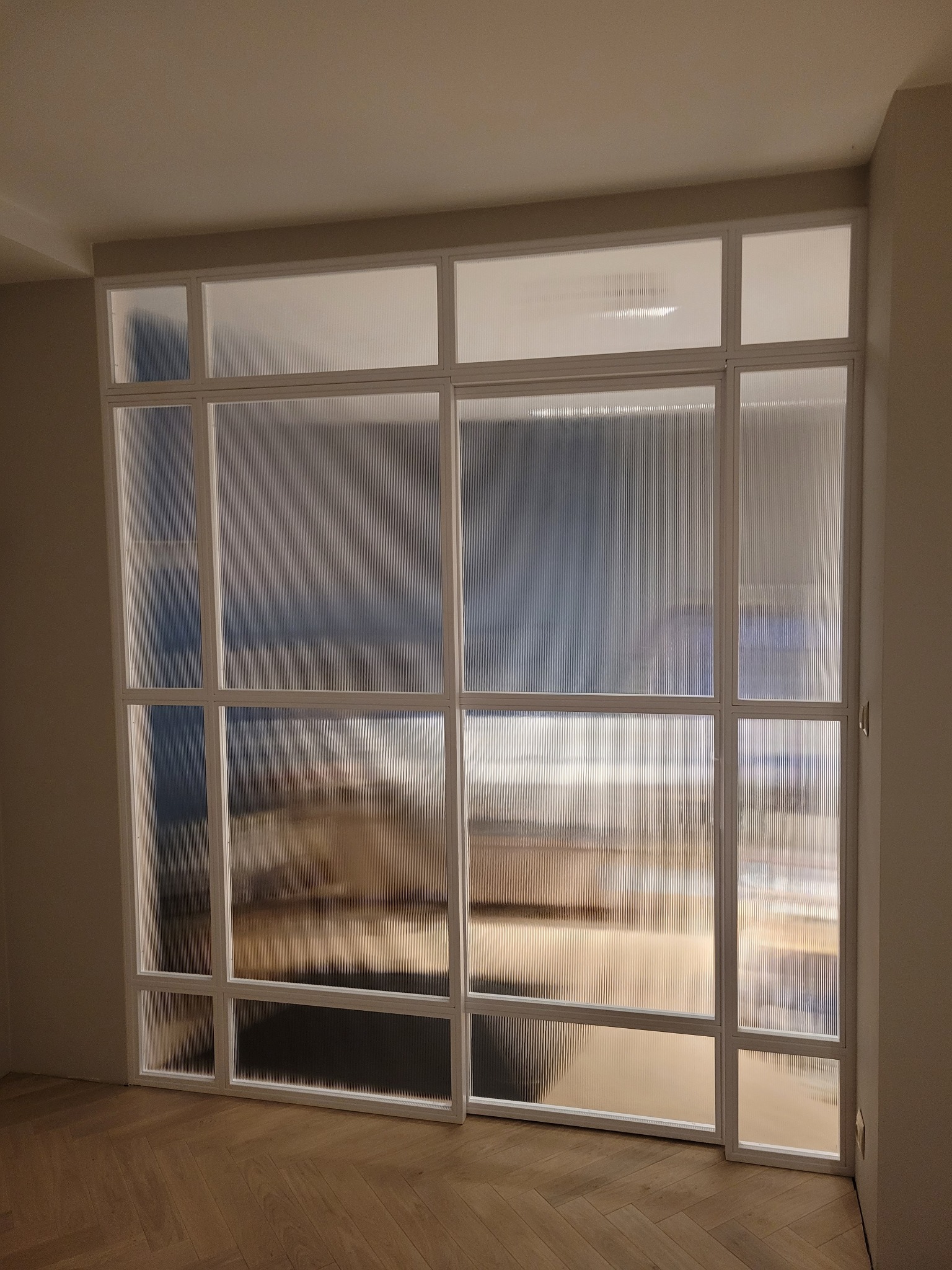 Nowoczesna, biała ścianka działowa z ryflowanego szkła, dzieląca przestrzeń w mieszkaniu, widoczna na tle parkietu ułożonego w jodełkę.