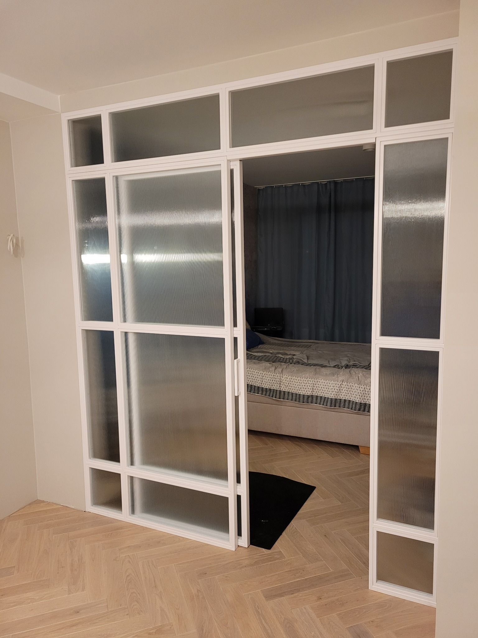 Nowoczesne, białe drzwi z przeszkleniami o fakturze lnu, oddzielające pokój z parkietem w jodełkę od sypialni z zasłoniętym oknem.