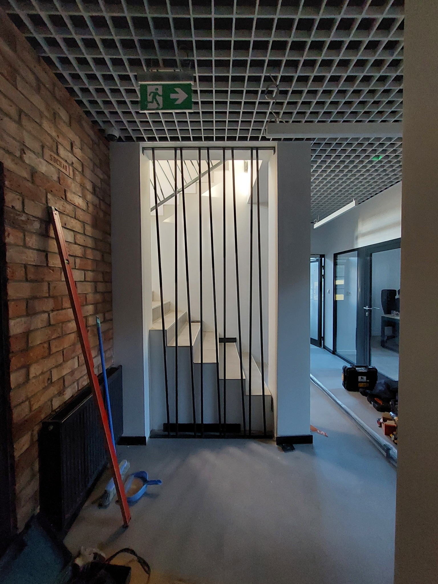 Wnętrze budynku w trakcie wykończenia: schody oddzielone pionowymi, czarnymi prętami, ceglana ściana po lewej, narzędzia i skrzynka narzędziowa na podłodze, sufit rastrowy z oznaczeniem drogi...