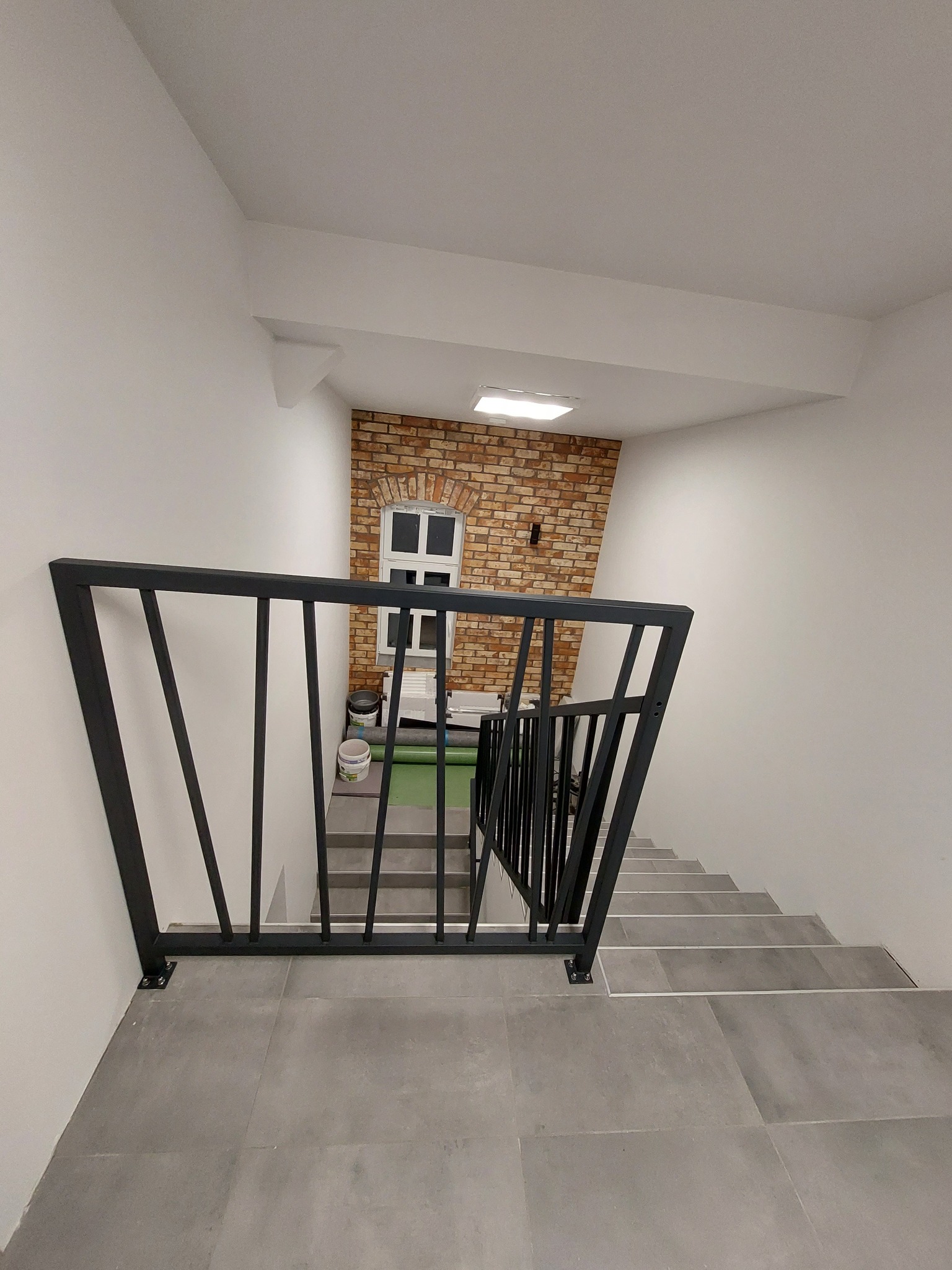 Metalowa balustrada schodowa w nowoczesnym wnętrzu z ceglaną ścianą w tle, widok z góry na szare stopnie i czarną konstrukcję.