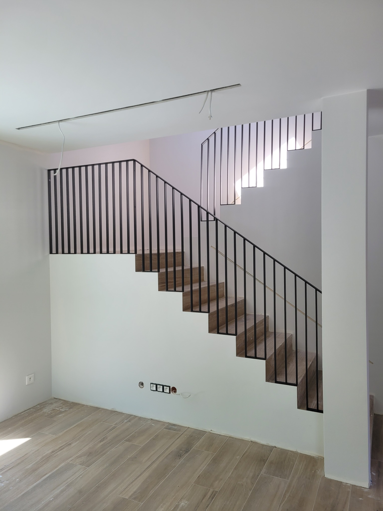 Nowoczesne schody z drewnianymi stopniami i czarną, minimalistyczną balustradą z pionowych prętów, widoczne gniazdka elektryczne i niedokończone prace wykończeniowe w tle.
