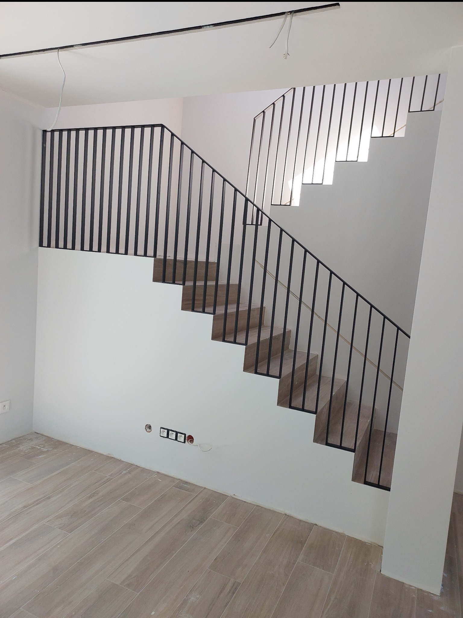 Nowoczesne schody wewnętrzne z czarną metalową balustradą, drewnopodobne stopnie, widoczne gniazdka elektryczne w ścianie, surowy stan wykończenia.