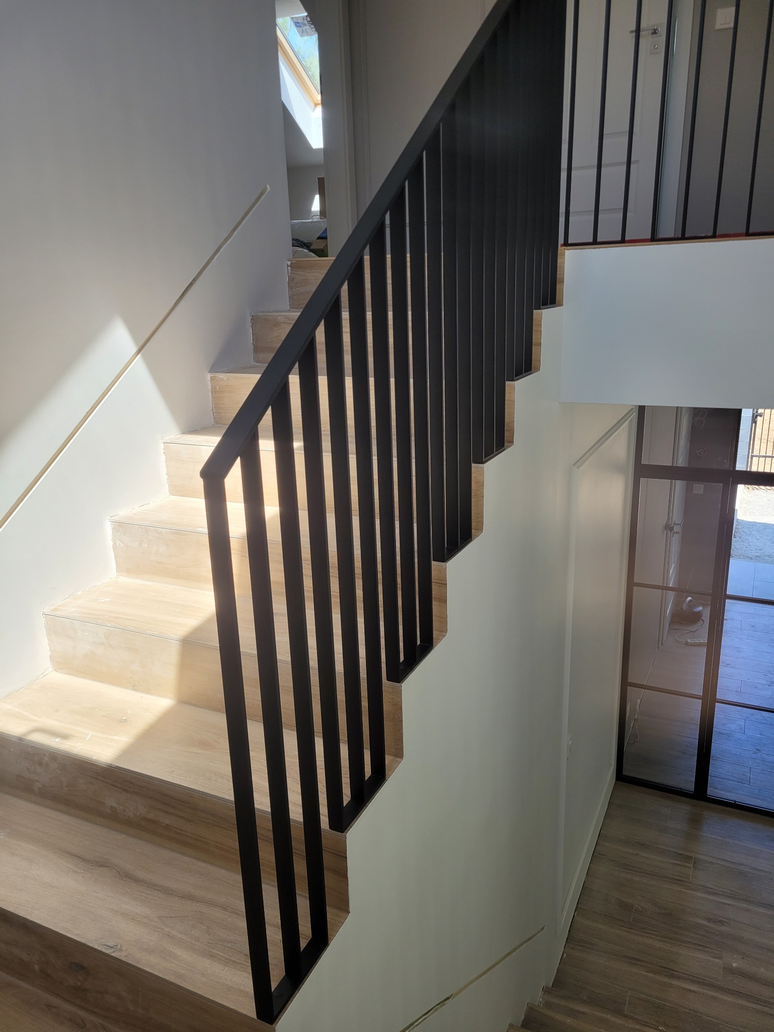 Nowoczesne schody wewnętrzne z drewnopodobnymi stopniami i czarną, minimalistyczną balustradą z pionowych prętów; widok z góry na dół, z oknem dachowym w tle.