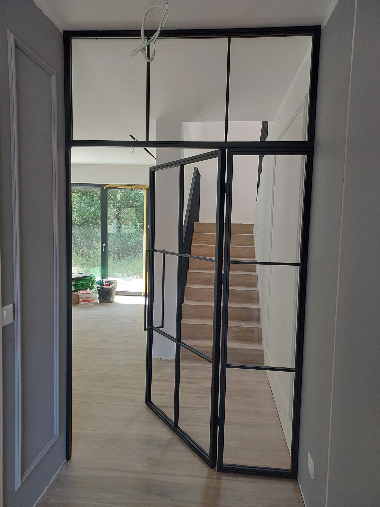 Szklane drzwi z czarną metalową ramą w stylu loftowym, widok na schody z drewnianymi stopniami i jasne wnętrze z widokiem na ogród.