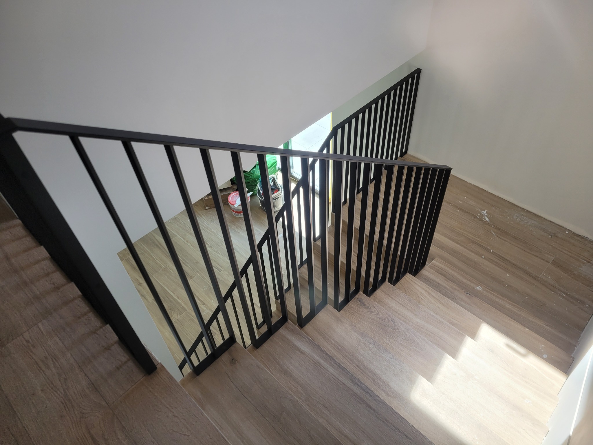 Widok z góry na drewniane schody z czarną, metalową poręczą z pionowych prętów, widoczne ślady po remoncie na podłodze i wiadro z farbą w tle.
