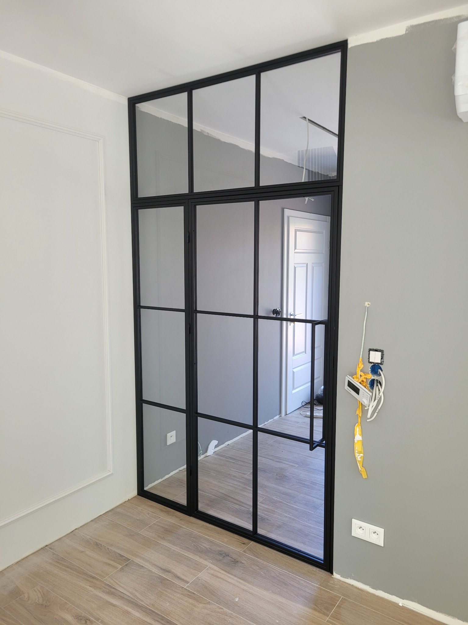 Dwuskrzydłowe drzwi wewnętrzne z czarnymi szprosami i szybami, widoczne kable elektryczne wystające ze ściany, niedokończone malowanie ściany na szaro.