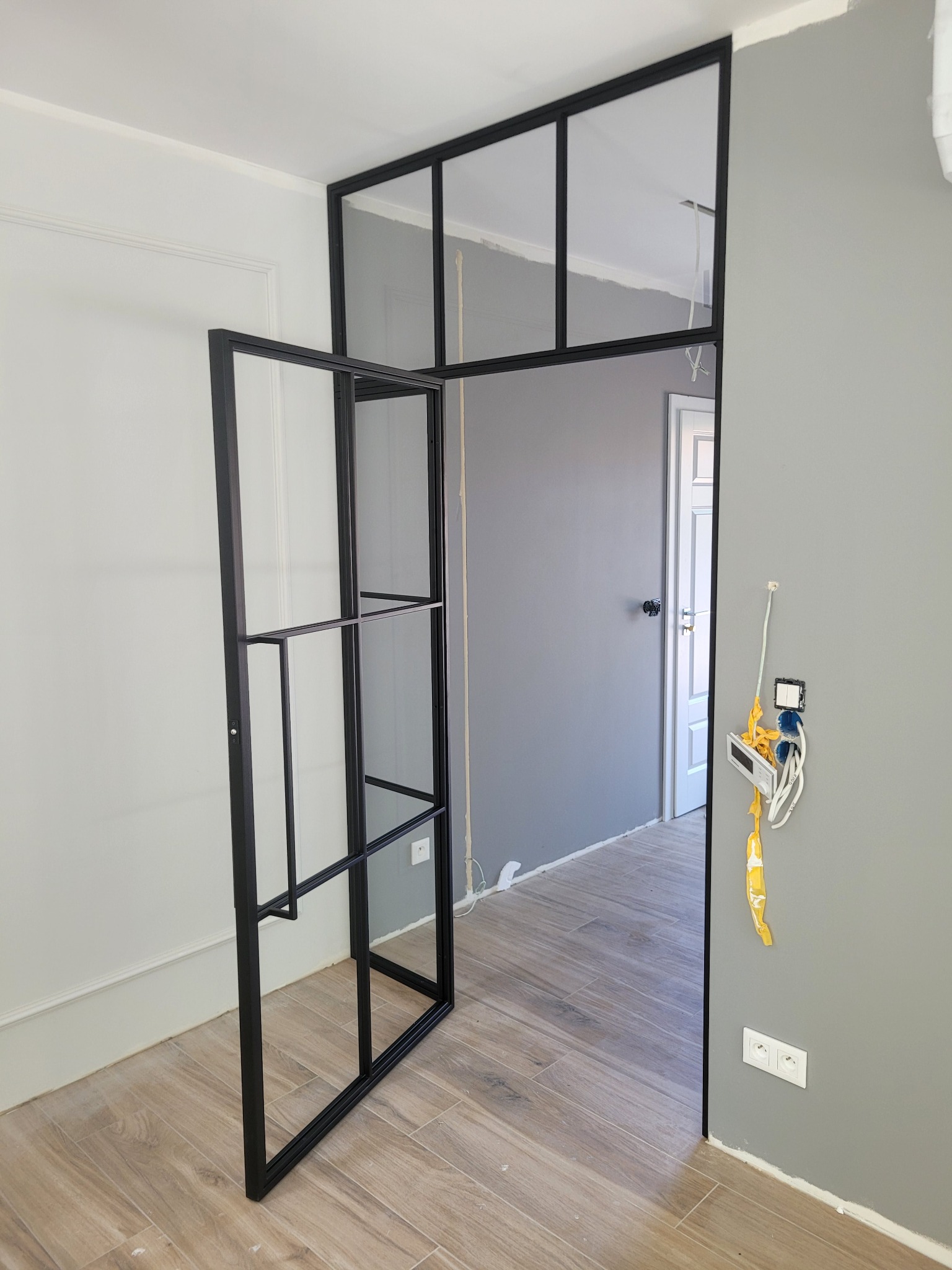 Metalowa konstrukcja z szybami w stylu industrialnym, tworząca ściankę działową i drzwi, w trakcie montażu; widoczne niedokończone prace elektryczne na ścianie.