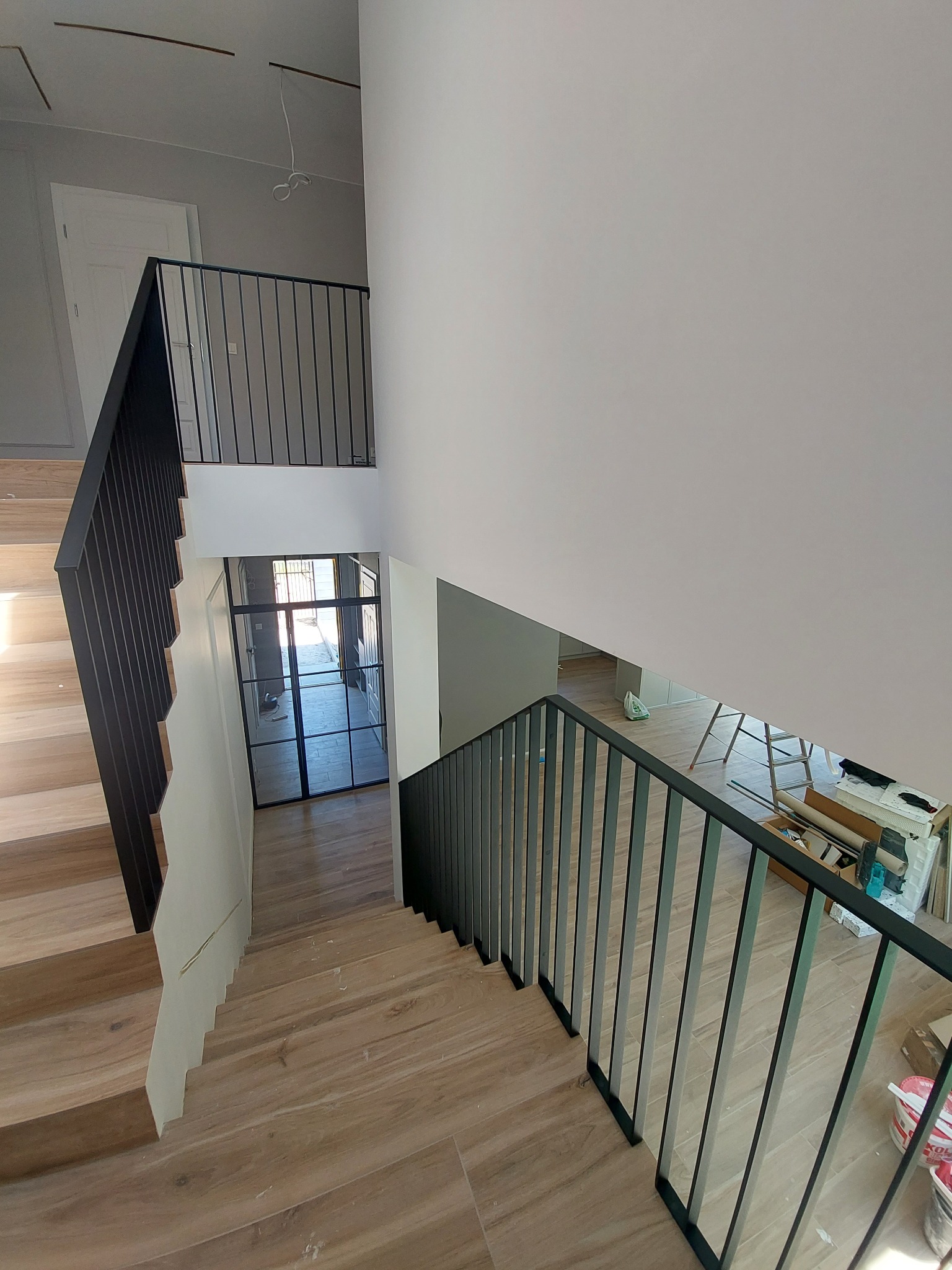 Nowoczesne schody z drewnopodobnymi płytkami i czarną metalową balustradą, widok z góry na dwupoziomowy hol z przeszklonymi drzwiami, w tle widoczne narzędzia i drabina.