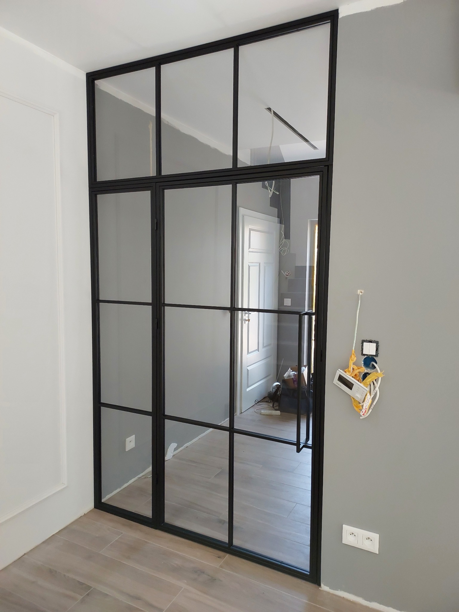 Nowoczesne, loftowe drzwi wewnętrzne z czarnymi szprosami i przezroczystym szkłem, widok na wnętrze z drewnianą podłogą i białą ścianą, instalacja elektryczna w trakcie wykończenia na szarej ścianie.