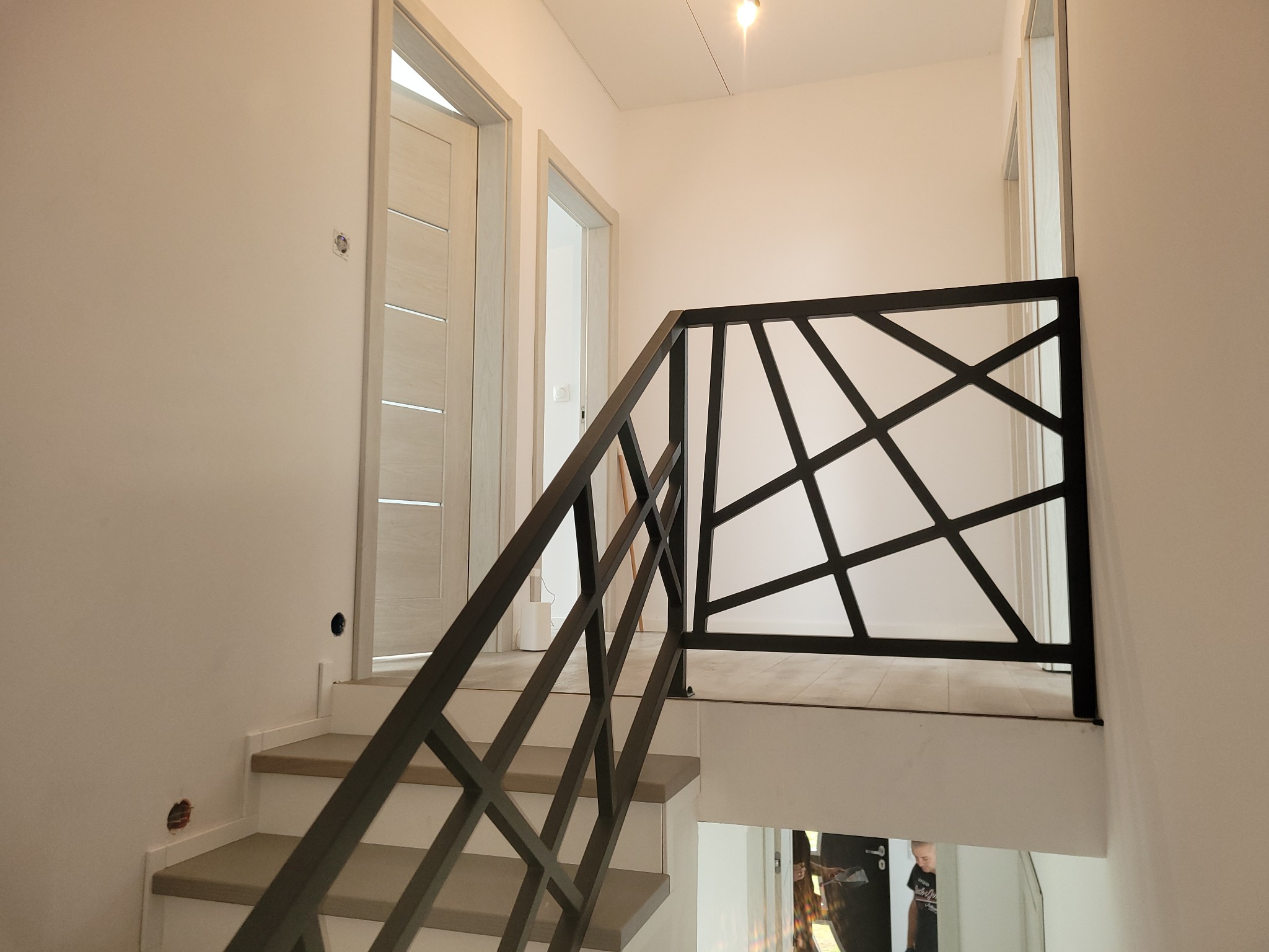 Nowoczesna, czarna balustrada metalowa o geometrycznym wzorze na schodach w jasnym wnętrzu, widoczne drzwi w tle.