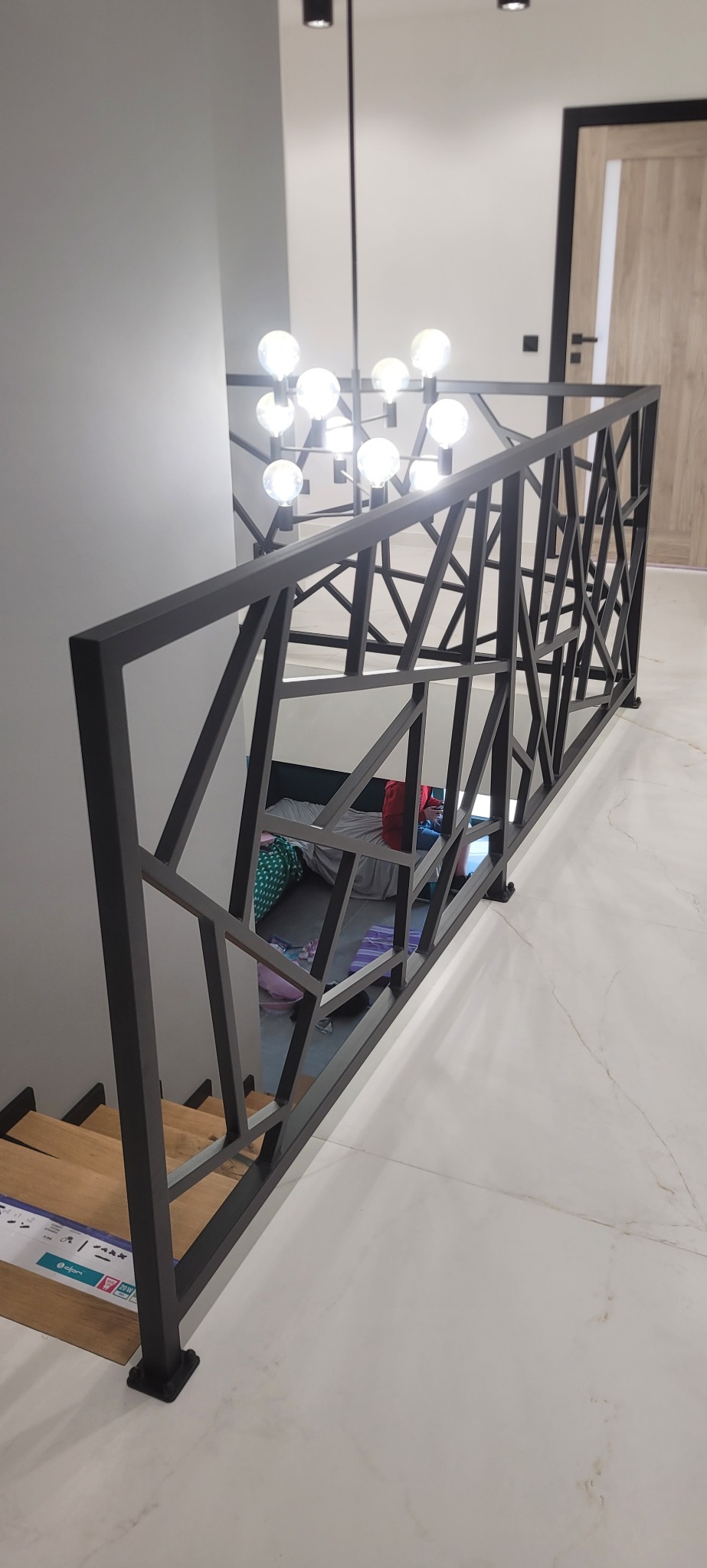 Nowoczesna, czarna balustrada schodowa z geometrycznym wzorem, widok z góry na drewniane stopnie i marmurową podłogę, w tle widoczny pokój z łóżkiem i lampa sufitowa w stylu industrialnym.