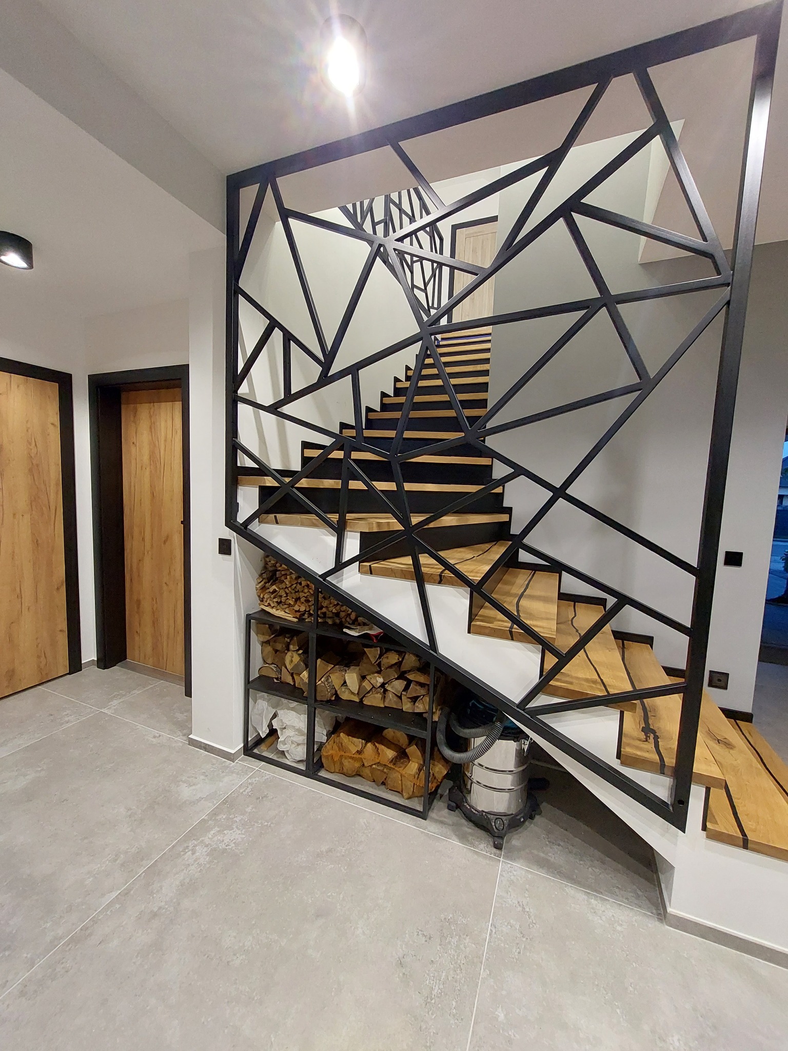 Nowoczesne schody z drewnianymi stopniami i geometryczną, czarną balustradą, pod schodami półka z drewnem na opał, obok odkurzacz przemysłowy.