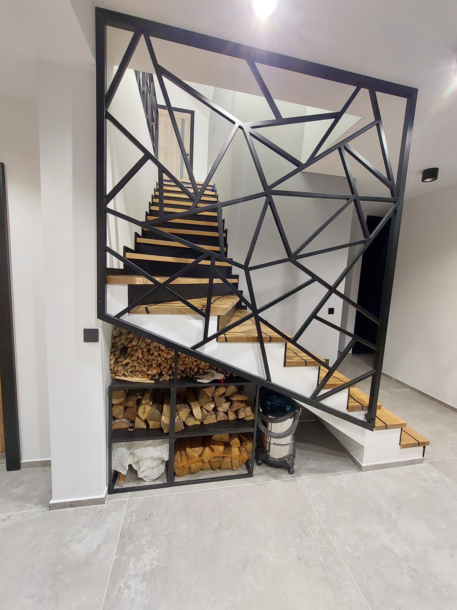 Nowoczesne schody z drewnianymi stopniami i ażurową metalową konstrukcją, pod schodami półki z drewnem opałowym, widoczny odkurzacz przemysłowy.
