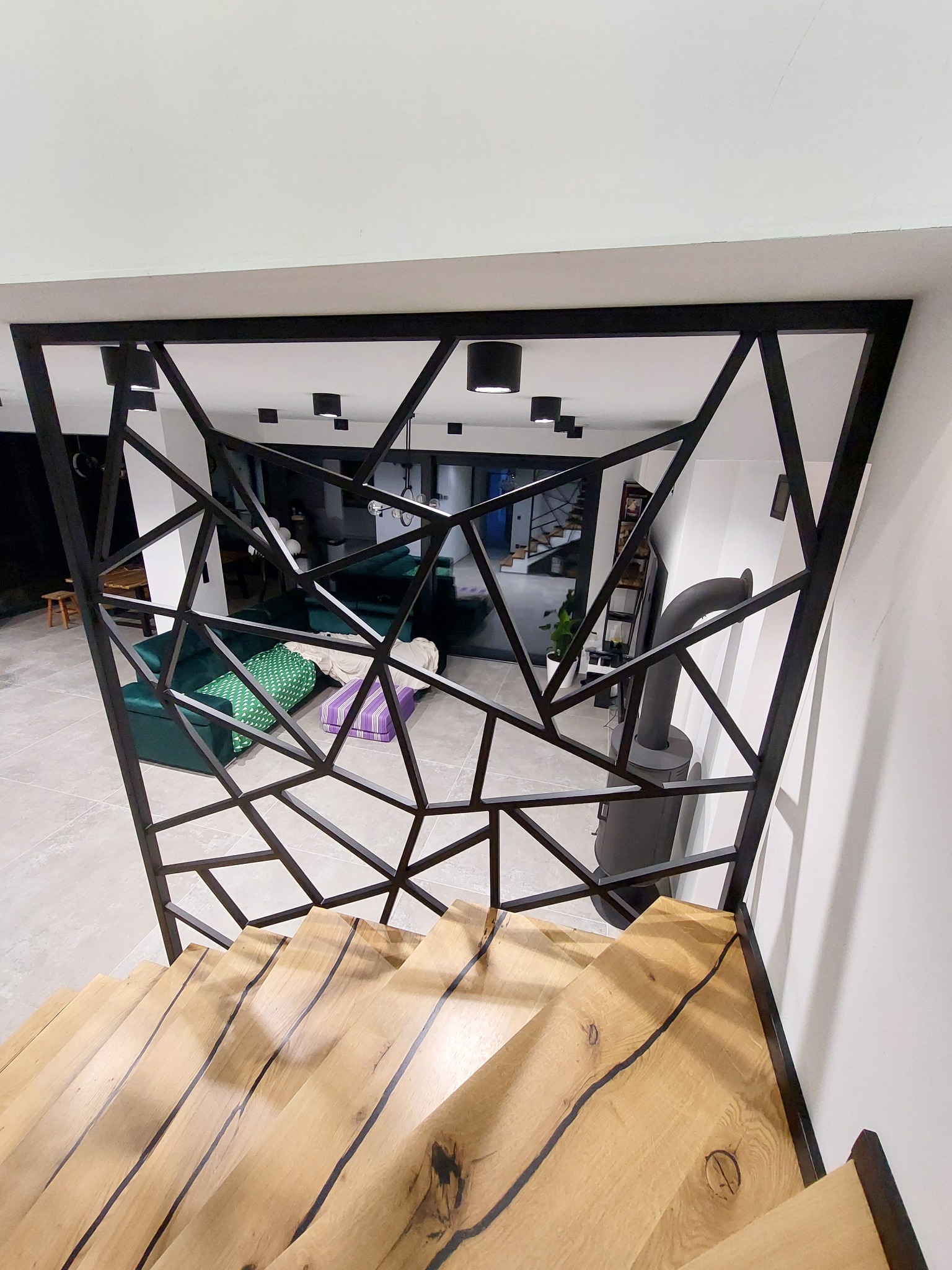 Nowoczesna, czarna konstrukcja stalowa o geometrycznym wzorze, pełniąca funkcję balustrady schodowej, z widokiem na salon z zieloną sofą i kominkiem.