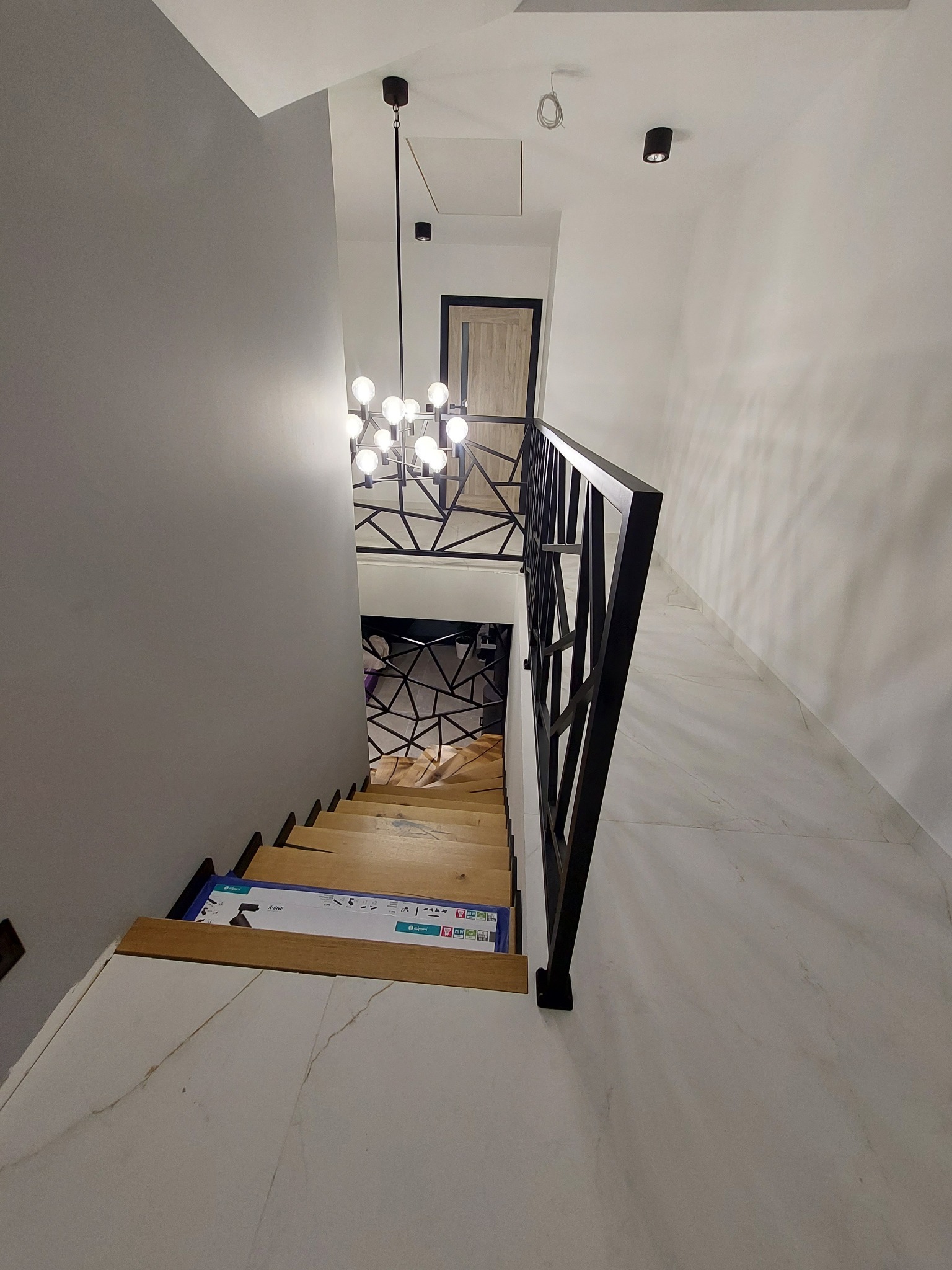 Klatka schodowa z drewnianymi schodami i czarną, geometryczną balustradą, widok z góry, z pudełkiem na schodach, nowoczesne oświetlenie.