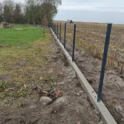 Betonowa podmurówka z zamontowanymi, czarnymi słupkami ogrodzeniowymi, widok na pole i drzewa w tle, pochmurny dzień.