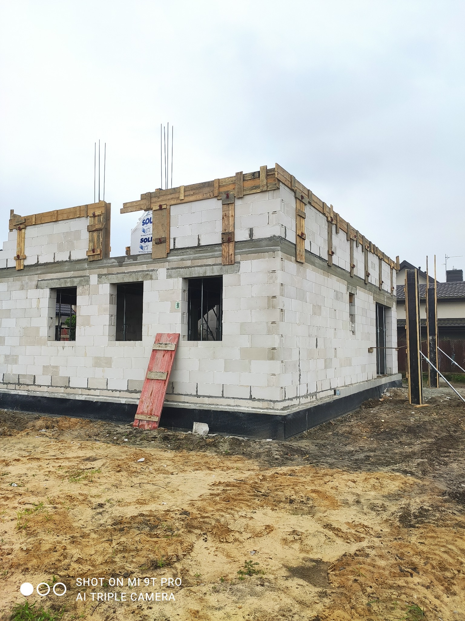 Surowy stan domu jednorodzinnego w trakcie budowy: białe bloczki, drewniane szalunki, wystające pręty zbrojeniowe, widoczne otwory okienne i drzwiowe.