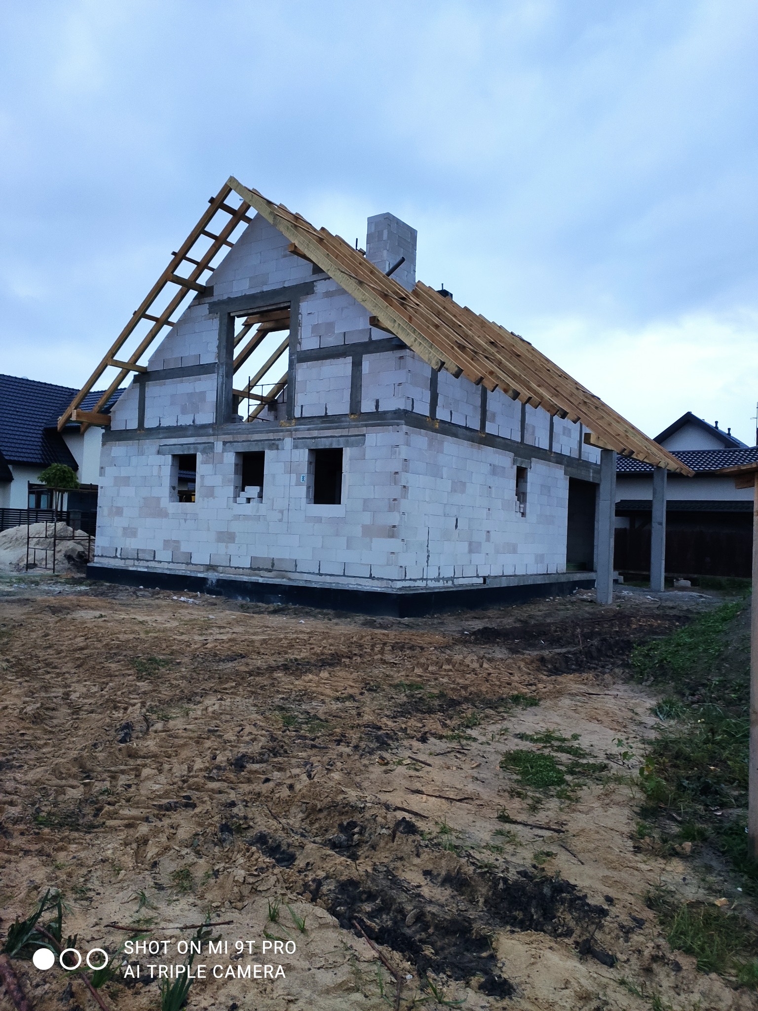 Surowy stan budowy domu jednorodzinnego z białych bloczków, widoczne drewniane elementy konstrukcji dachu, na zewnątrz teren budowy z piaskiem i błotem.