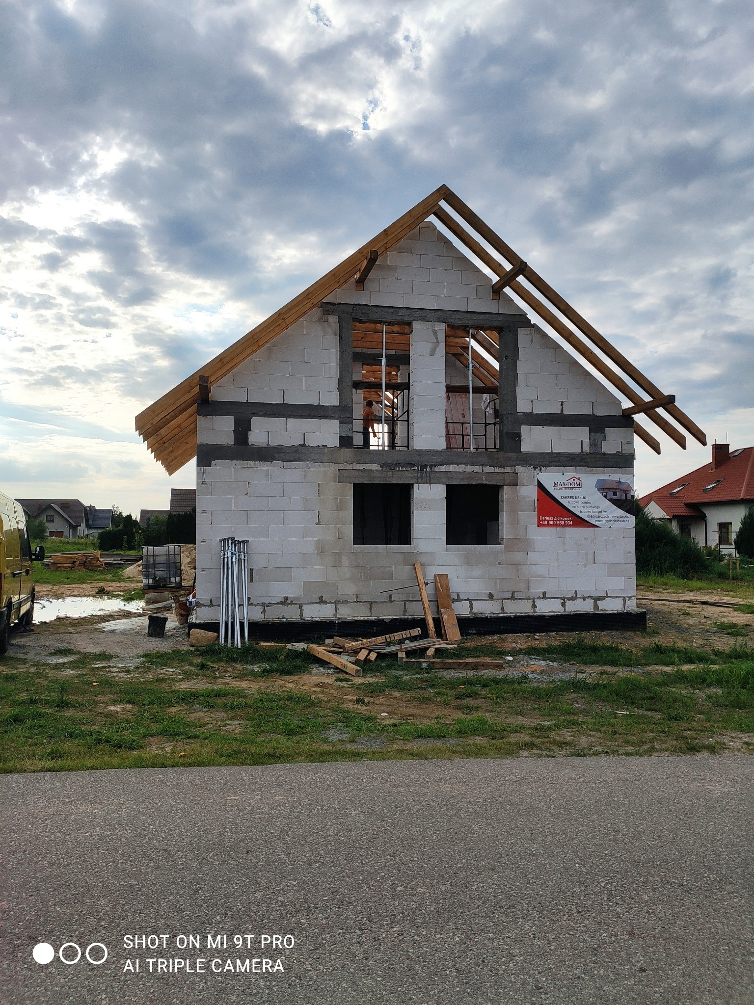 Budowa domu jednorodzinnego w stanie surowym otwartym, widoczne ściany z bloczków, drewniana konstrukcja dachu, tablica reklamowa firmy budowlanej na ścianie, teren budowy z kałużami i materiałami...