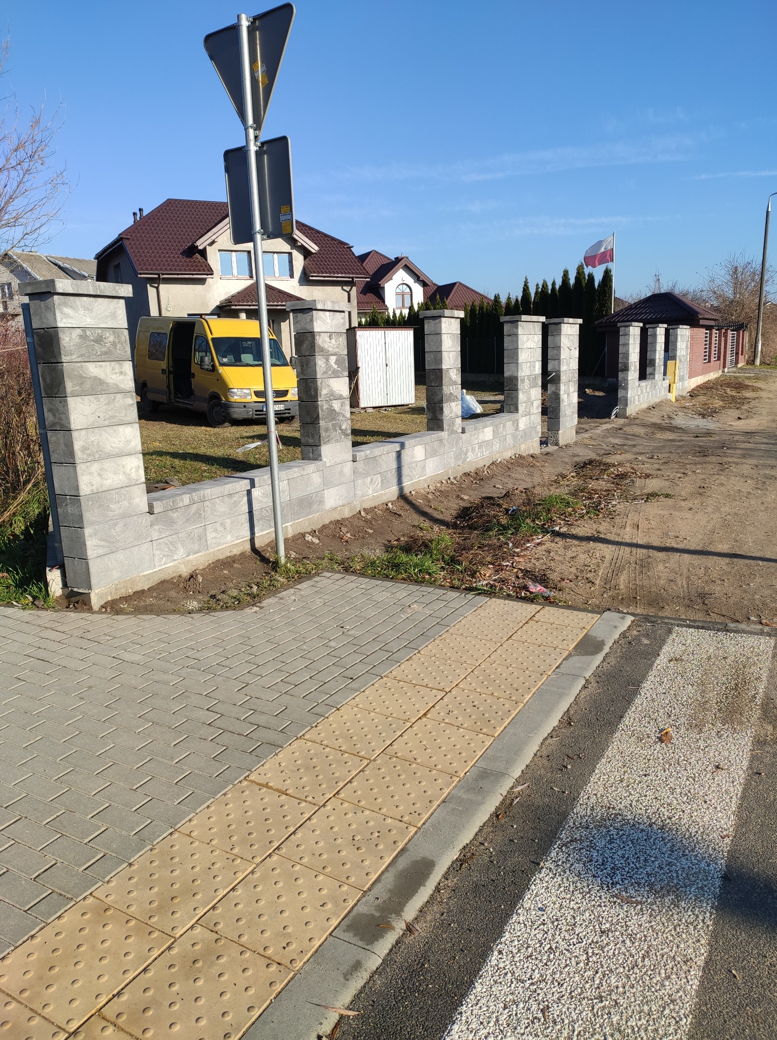 Wznoszony z szarych bloczków betonowych płot na posesji z żółtym busem w tle, widok z chodnika dla pieszych.