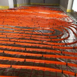 Pomarańczowe rury ogrzewania podłogowego ułożone w pętle na folii izolacyjnej z siatką w surowym wnętrzu budynku w trakcie budowy.