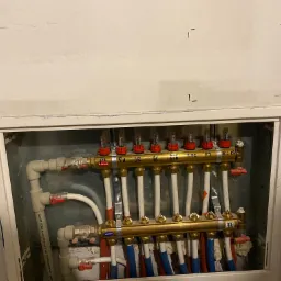 Rozdzielacz ogrzewania podłogowego w zabudowie ściennej z widocznymi rurami doprowadzającymi wodę, z oznaczeniami na górnej listwie i zaworami regulacyjnymi.