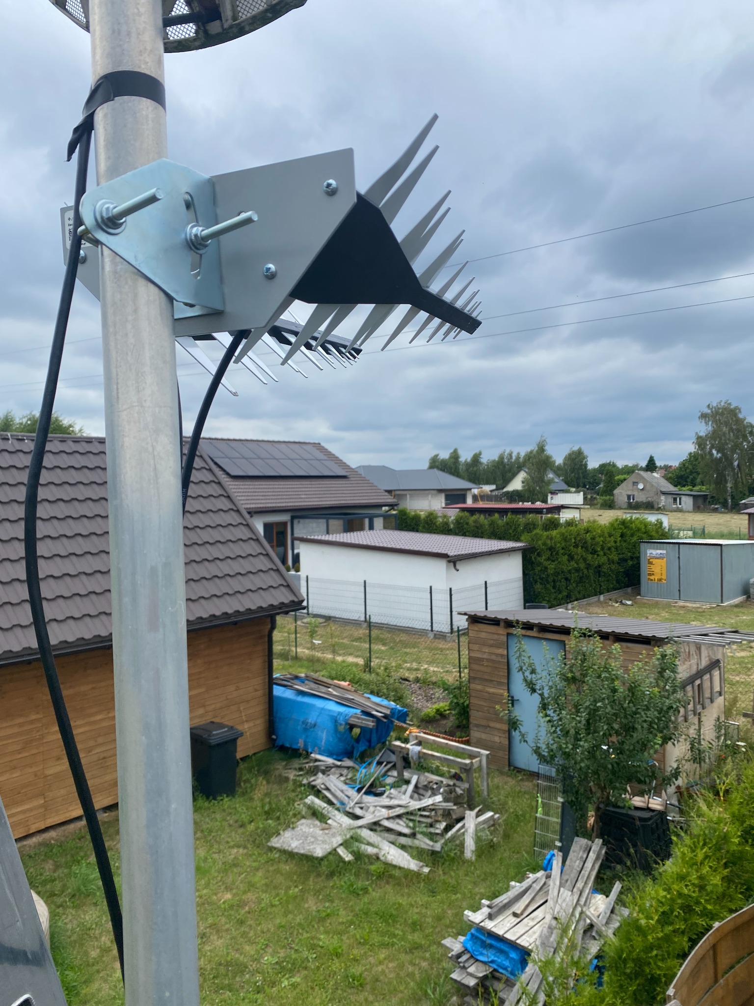 Antena szerokopasmowa typu Yagi-Uda zamontowana na metalowym maszcie, widok z góry na zabudowę jednorodzinną i pochmurne niebo.