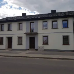 Dwupiętrowy budynek po renowacji elewacji, z kremową fasadą, szarymi narożnikami i ciemnymi oknami, sfotografowany z poziomu ulicy w pochmurny dzień.