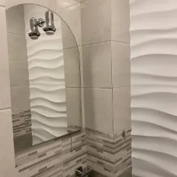 Nowoczesna łazienka z beżowymi płytkami, dekoracyjną ścianą z falistym wzorem i owalnym lustrem z oświetleniem.