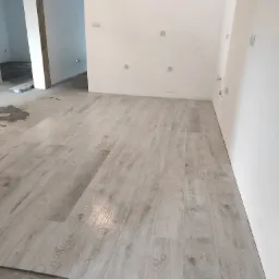 Wnętrze w trakcie prac wykończeniowych; ułożona podłoga z paneli imitujących drewno w odcieniach szarości, widoczne surowe ściany z wystającymi przewodami elektrycznymi oraz fragmenty nieukończonej...