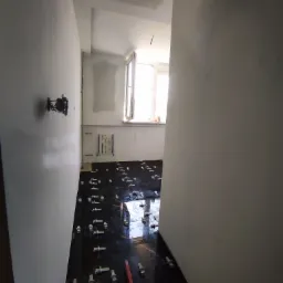 Proces układania ciemnych, błyszczących płytek podłogowych z użyciem krzyżyków dystansowych, widoczny fragment ściany z wystającymi przewodami elektrycznymi i okno w tle.