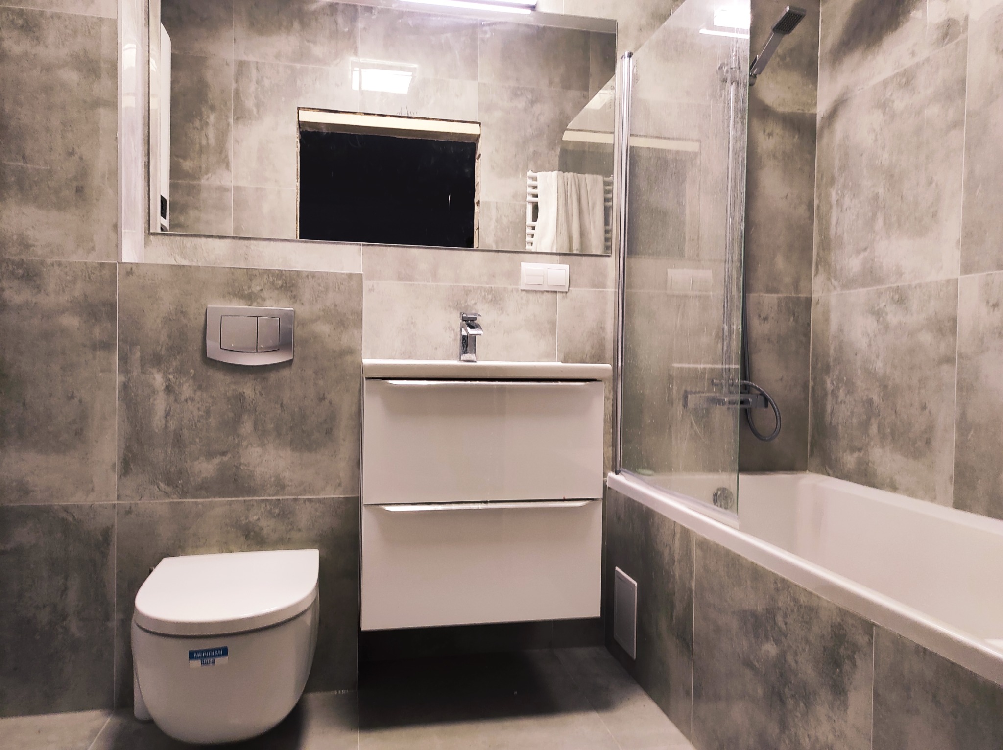 Nowoczesna łazienka z szarymi płytkami imitującymi beton, białą szafką pod umywalką z chromowaną baterią, wiszącą toaletą i wanną za szklanym panelem.