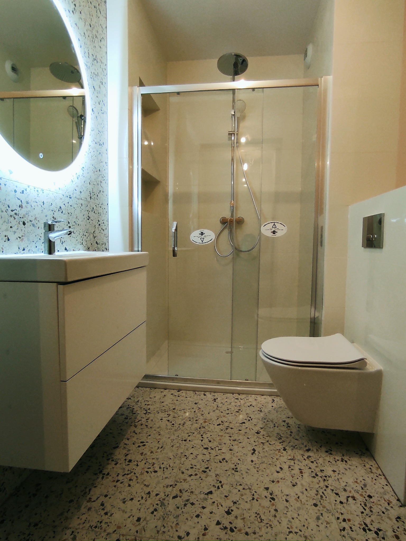 Nowoczesna łazienka z kabiną prysznicową, wiszącą toaletą i umywalką z szafką, wykończona płytkami lastryko na ścianie i podłodze, oświetlona lustrem LED.