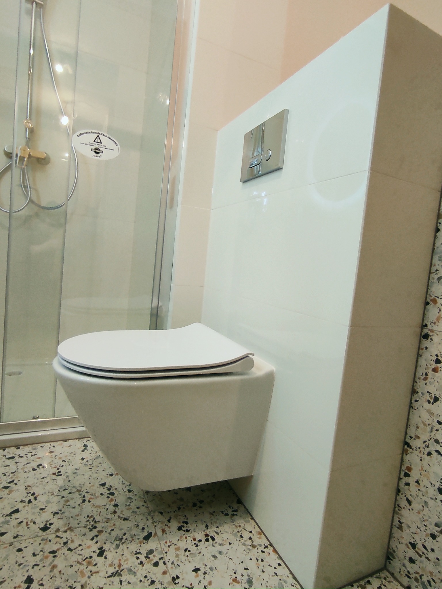 Wisząca toaleta z podtynkową spłuczką, obok kabina prysznicowa z naklejką ostrzegawczą, na podłodze płytki lastryko.