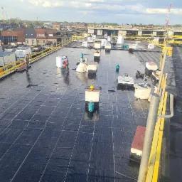 Widok z góry na płaski dach budynku komercyjnego w trakcie hydroizolacji, z widocznymi rolkami papy, butlami gazowymi i pracownikami w oddali, na tle zabudowy miejskiej.