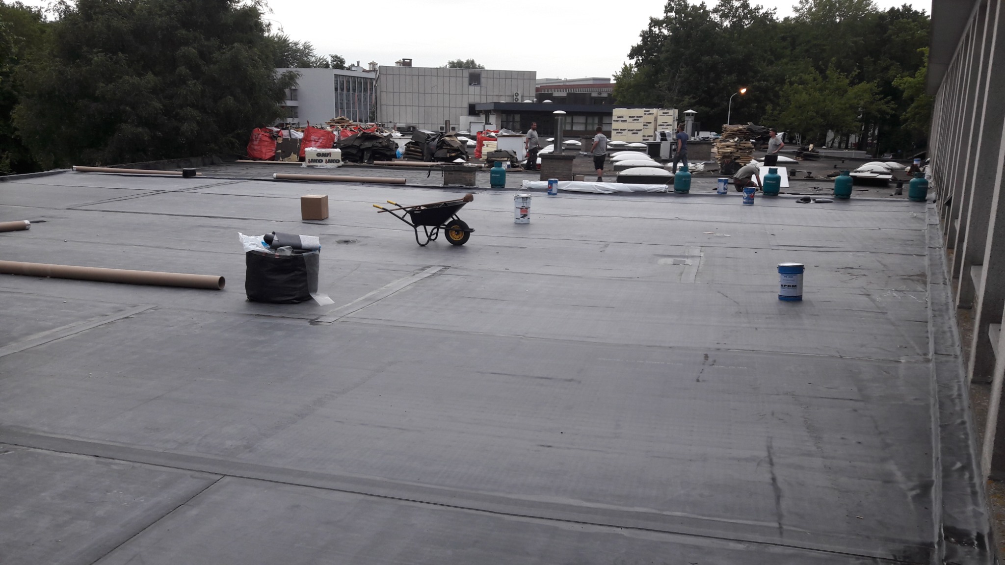 Montaż membrany EPDM na płaskim dachu budynku usługowego. Widoczne rolki membrany, wiaderka z klejem, taczka i pracownicy w tle.