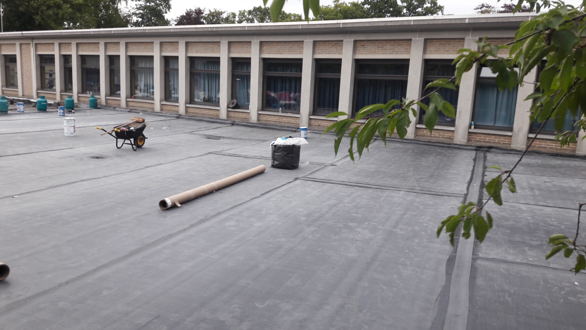 Bitumiczna powierzchnia dachu budynku z widocznymi szwami, w tle wózek z narzędziami, pojemniki z farbą, worki na śmieci i budynek z oknami, ujęcie z góry z gałęzią drzewa w prawym rogu.