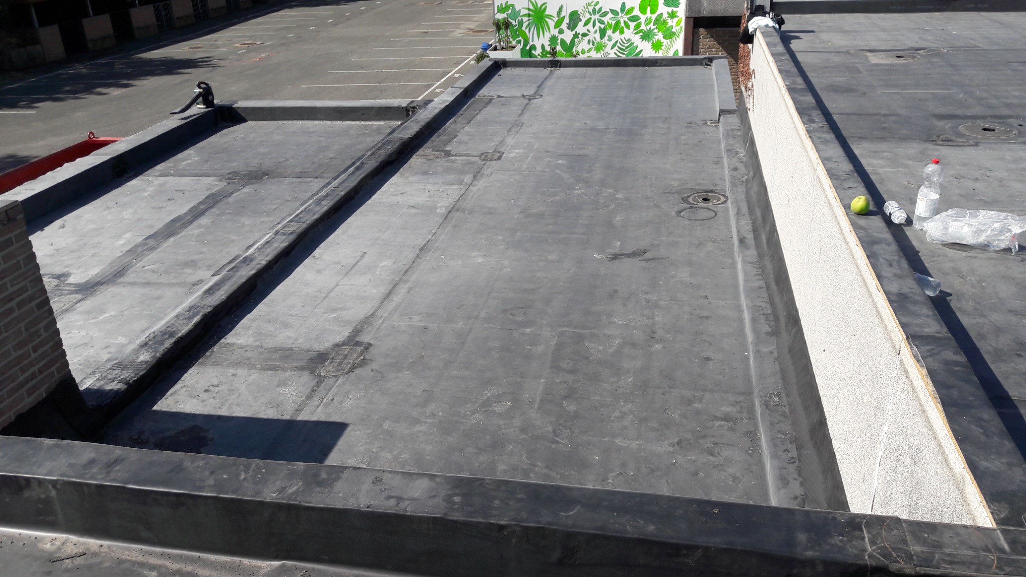 Płaski dach pokryty papą termozgrzewalną, widoczne łączenia i detale wykończeniowe, na dachu leżą butelki z wodą i jabłko, w tle parking.