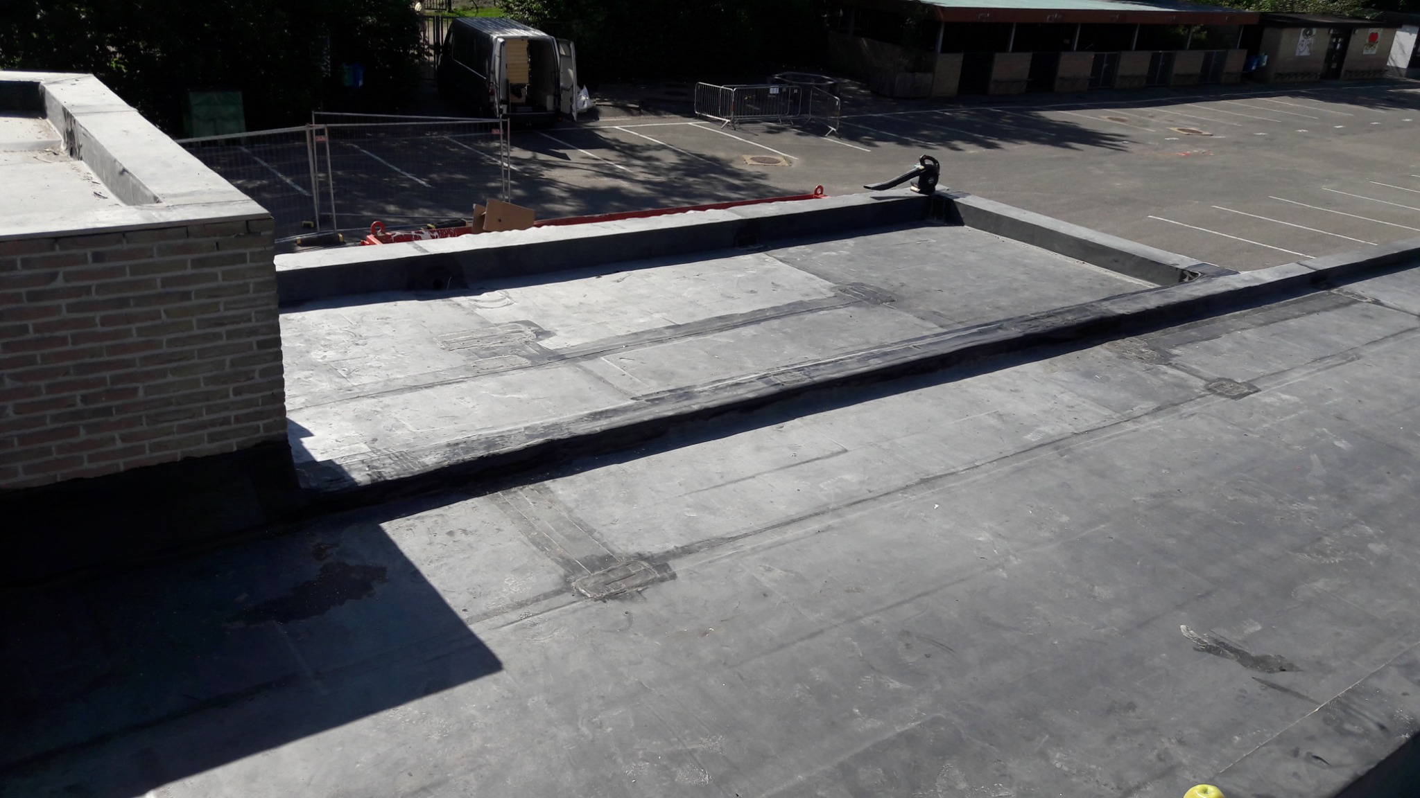 Płaski dach pokryty papą z widocznymi miejscami łączeń i napraw, w tle fragment parkingu i budynków.