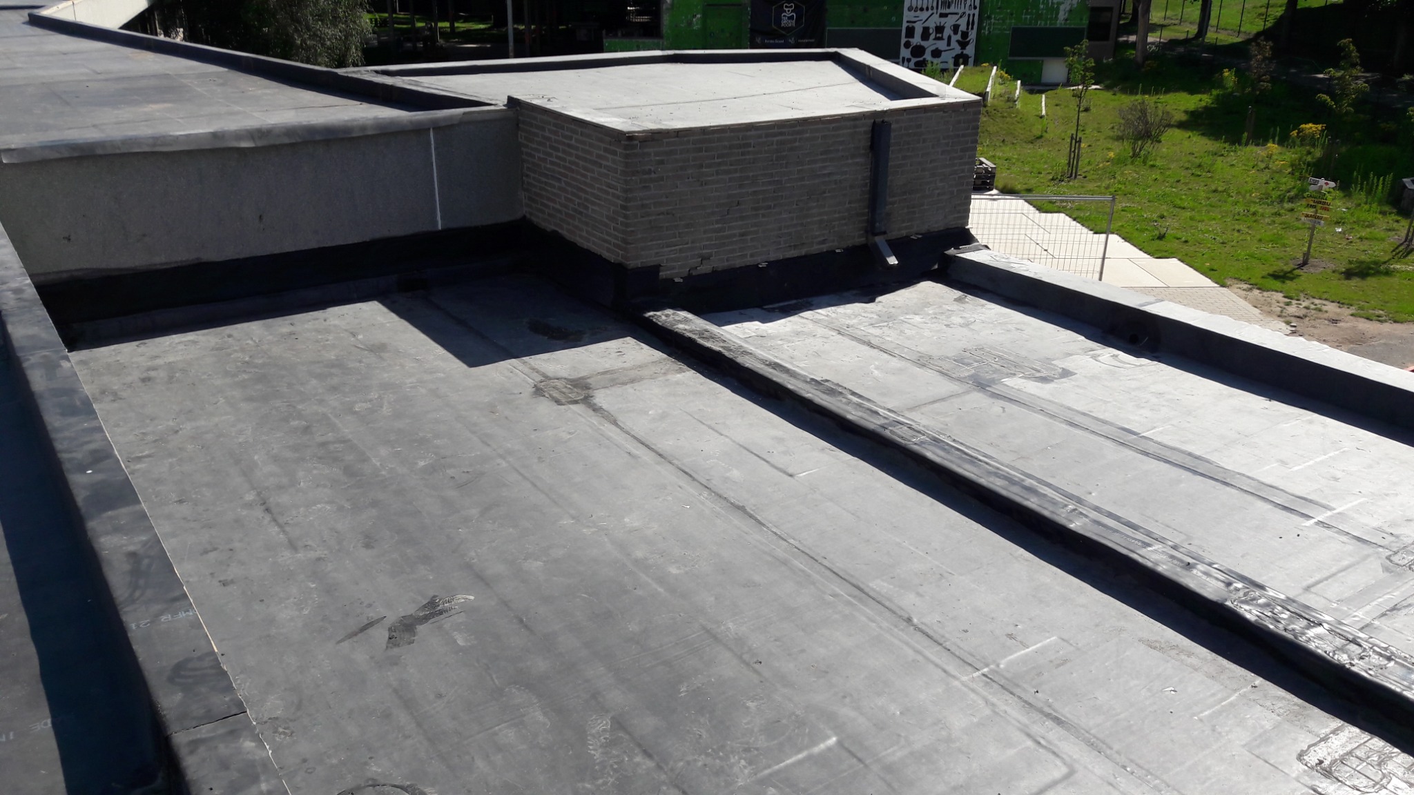 Płaski dach pokryty papą z widocznymi łączeniami i kominem wentylacyjnym z cegły, na tle zieleni.