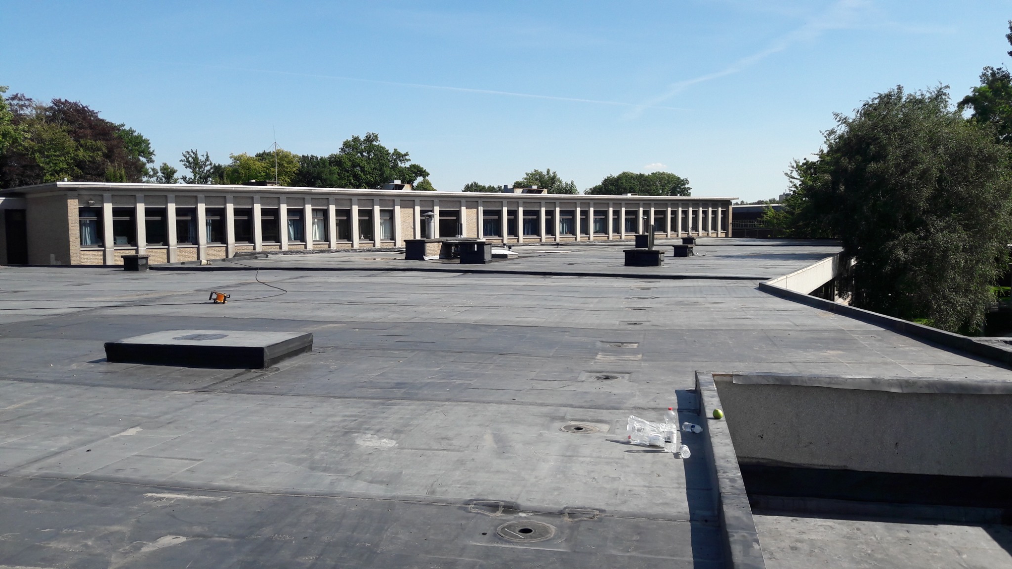 Płaski dach budynku usługowego pokryty papą, widoczne kominki wentylacyjne, narzędzie dekarskie oraz śmieci, w tle budynek z oknami.