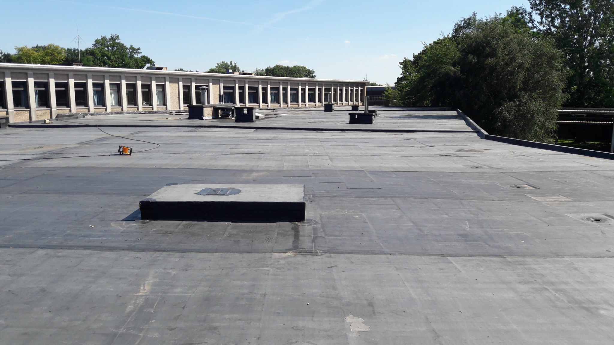 Płaski dach pokryty papą termozgrzewalną z widocznymi kominkami wentylacyjnymi i urządzeniem do spawania papy w tle, budynek w tle.